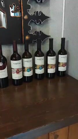 The video shows a row of wine bottles lined up on a wooden surface, likely a bar or counter. Behind the bottles, a mirror reflects a person wearing an orange shirt and holding a phone, possibly taking a picture or video. The mirror is framed by a decorative element with a black background and white outlines of stylized shapes, possibly bats or leaves. The bottles are of various colors and labels, suggesting different types of wine. The lighting appears to be artificial, coming from above. The scene is indoors, with brickwork visible in the background reflection.