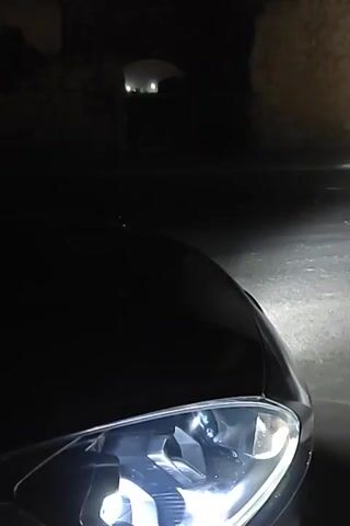 The video shows a car at night. The camera is positioned near the front of the car, focusing on the headlight and the road illuminated by it. The background includes a dark structure, possibly a building or archway, followed by a road with road signs, a streetlight, and other distant lights. The road appears wet or reflective. The camera pans slightly, following the path of the light.