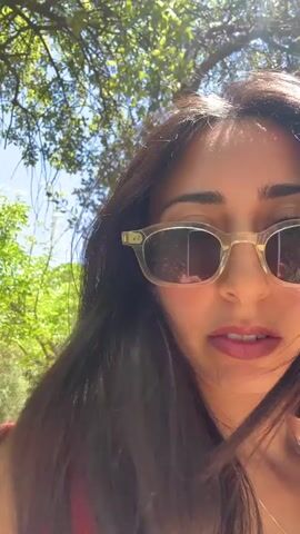 This is a selfie video of a young woman with dark hair and sunglasses, taken outdoors on a sunny day. She is wearing a red top. The background is blurred but appears to be a natural setting with green trees and blue sky, suggesting it's daytime. The woman is looking directly at the camera and her lips are slightly parted. There is no visible text in the media. The context provided indicates the location is Al-Eizariya, Palestinian Territories.
