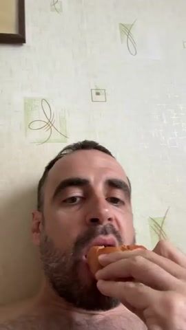 A middle-aged man with a beard is seen from a low-angle, close-up shot, taking a bite out of a small, baked item. The man has short, dark hair and is shirtless. He is holding the baked good with his right hand. His face is expressive, and he appears to be tasting the food intently. The background is a wall with a patterned wallpaper. A small, framed picture hangs on the wall in the upper-left corner of the frame.