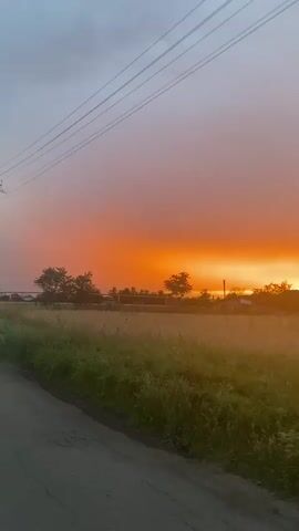 This video captures a picturesque sunset over a rural landscape in Yutin, Russia. The scene is dominated by the vibrant hues of the setting sun, which paints the sky in shades of orange, pink, and purple. A dramatic contrast is created by the dark grey clouds that loom above, hinting at recent or approaching rain.

In the foreground, a narrow asphalt road runs along the left side of the frame, bordered by lush green grass and tall weeds. Beyond the road, a field of dry, golden crops stretches across the middle ground, its texture appearing soft and slightly blurred. Silhouetted against the sunset are a few scattered trees and the outlines of buildings, suggesting a small village or farmsteads. Power lines are visible in the upper portion of the sky, adding a touch of human infrastructure to the natural beauty.

The overall mood is serene and tranquil, with the dramatic sky evoking a sense of awe and the quiet countryside suggesting peace. The time of day is clearly sunset, characterized by the intense colors and long shadows, though specific details about the weather beyond the clouds are not explicitly visible. There is no visible text in the video.