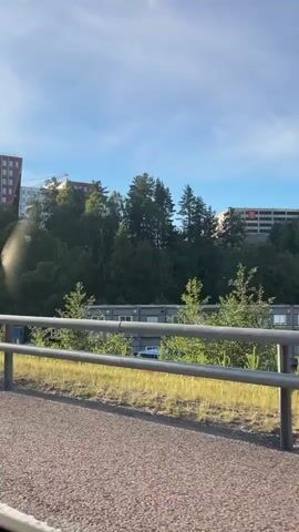 The video shows a view from inside a moving vehicle, likely a car. The perspective is from the passenger side, looking out at a semi-urban or suburban landscape. In the foreground, a grey gravel surface is visible, possibly a road or pathway, bordered by a metal guardrail with grey posts and horizontal bars. Beyond the guardrail, dry yellow grass and some green vegetation are present. Further in the background, a row of modular buildings, appearing to be temporary structures or portable offices, are lined up. One building has the letters "HAB" on it. Behind these buildings, a dense green forest covers a hillside. On top of the hill, several multi-story buildings, some appearing to be under construction with cranes visible, are situated amongst the trees. The sky is blue with some scattered clouds, and the overall lighting suggests it is daytime, likely late afternoon or early morning due to the angle of the sun casting long shadows. The video captures the motion of the vehicle as it passes by the scenery.