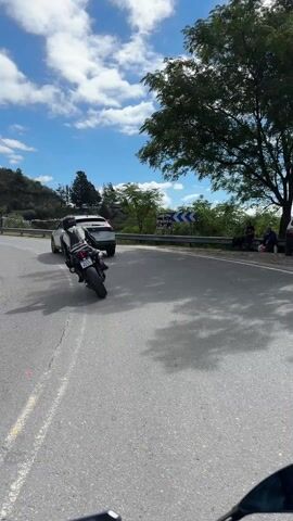 The video shows a motorcyclist, dressed in full black and white gear, riding along a paved road, followed by a white SUV. The road winds around a hill covered in trees and rocks. The sky is blue with scattered white clouds. A road sign decorated with colorful pennants stands on the side of the road.