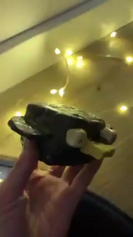 A hand is holding a dark, handmade clay figure of a bird. The bird is boxy with a yellow beak and white eyes. It has stubby wings. There are yellow fairy lights in the background. The hand rotates the figure to show it from different angles. The background consists of a light-colored wall and a wood floor.