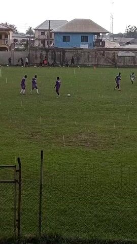 The video shows a soccer game in progress on a green field. It is raining, and droplets are visible. The soccer players are wearing different color uniforms: one team wears white and the other wears purple. There is a soccer ball in the center of the field with players from each team around it. In the background, behind a grey concrete wall, are buildings. There is a black chain-link fence in the foreground.