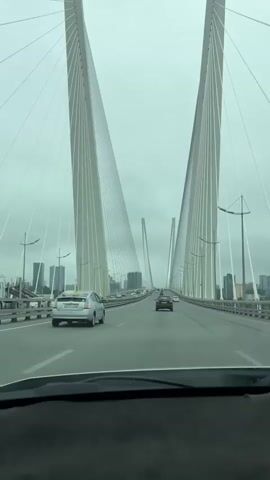 The video shows a view from the driver's seat of a car as it travels across a large cable-stayed bridge. The bridge has two massive white pylons that are very tall and lean inwards. Numerous white cables extend from the pylons to support the bridge deck. The sky is overcast and gray. Several cars are visible on the bridge ahead of the car. In the distance, a city skyline with buildings can be seen on both sides of the bridge. On the dashboard of the car, there are three gauges with circular dials. The car is moving forward on the bridge.