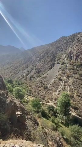 A video pans across a rugged, mountainous landscape under a clear blue sky. The terrain is steep and rocky, with sparse vegetation consisting of dry brush and scattered trees. A visible path or road winds its way up the mountainside, marked by tire tracks. The sunlight casts strong shadows, highlighting the textures of the rocks and the arid environment. The overall impression is one of a remote and untamed natural setting.