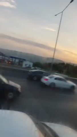 The video captures a view from inside a car driving on a highway. The sky is overcast with hues of orange and pink, indicating either sunrise or sunset. Several cars are visible on the highway, including a dark-colored minivan in the foreground, a white sedan, a silver hatchback, and another dark-colored sedan further back. The cars appear to be in traffic, moving at a slow pace. In the background, there are buildings that resemble a gas station with signs and pumps. Hills or mountains are visible in the distance. The camera is positioned at an angle, suggesting it is mounted on the dashboard or held by a passenger. The view is partially obscured by raindrops on the windshield. The overall scene suggests a typical highway commute during a potentially transitional time of day, with light traffic.
