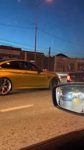 A driver, possibly stuck in traffic, films a yellow BMW M4 through their side window at dusk. The yellow car sits motionless on the driver's side, with its rear lights illuminated. A dark-colored vehicle in front blocks much of the view. The side mirror shows other cars, many with bright headlights, also stopped. A brown building behind a metal fence is visible. The sky is twilight blue, and streetlights illuminate the scene. Traffic signs are visible near the dark-colored vehicle.