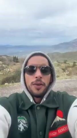 A man is recording a selfie video, standing on a dirt road surrounded by mountains and greenery. He is wearing sunglasses, a gray hoodie, and a green jacket with patches on it. He speaks directly to the camera. The background shows a cloudy sky, mountains in the distance, and scattered trees. As the camera moves slightly, a small building with blue accents becomes visible in the background.