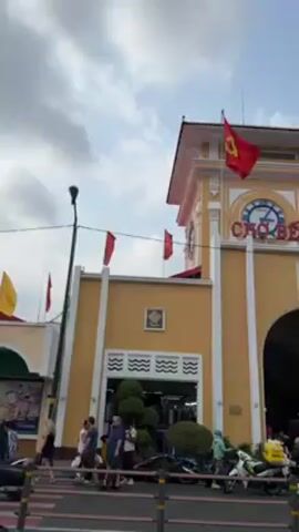 The video shows the exterior of the Ben Thanh Market in Ho Chi Minh City, Vietnam. The market building is painted a light yellow with white accents, and a red Vietnamese flag flies from the top of the building. The entrance to the market is an arched gateway. Several people are seen walking near the market entrance. Cars are also visible. The sky is blue with some clouds.