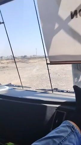 The video provides a view from inside the cabin of a large vehicle, possibly a truck or a bus, positioned in an outdoor, dusty environment. The interior of the cabin is visible, showcasing the steering wheel, dashboard, and side mirrors. Outside the windows, there is a view of a construction or industrial site with dirt mounds and some machinery. The overall scene suggests the vehicle is either stationary or moving slowly through this outdoor work area. The sky appears clear and blue.