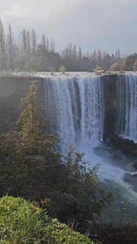 The video shows a scenic view of a wide waterfall cascading over a rocky edge into a river below.  There are trees and greenery visible on either side of the waterfall, as well as on the ledge above. The sky is overcast, creating soft, diffused light. The overall setting appears to be a natural park or wilderness area.