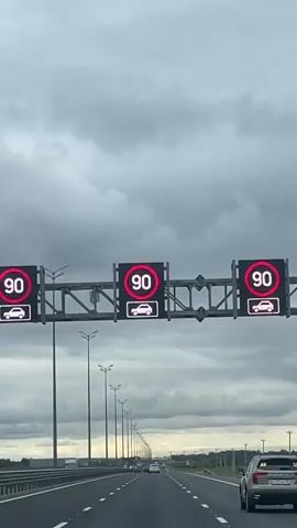 A speed limit sign displaying 90 and a car icon is visible overhead on a highway. The camera pans down to reveal a long stretch of highway with multiple lanes and a row of streetlights on the right side. The sky is overcast with dark clouds. Several cars are visible on the highway in the distance. The perspective suggests the video is being recorded from inside a moving vehicle.