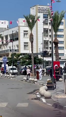 The video shows a street scene in front of a hotel. A white building with the words "DOME HOTEL" in large letters is visible. There are palm trees in front of the hotel. Turkish flags are strung across the street. Several people are visible on the street, including some in uniform. A white van is parked on the street. The sky is blue and the weather appears to be sunny.