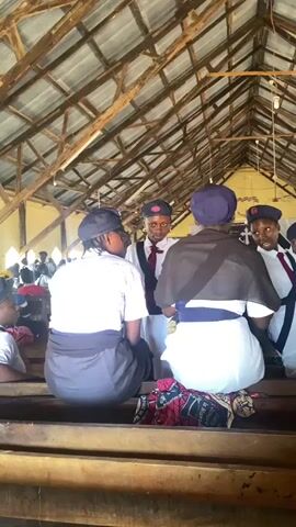 The video shows four individuals, presumably teenagers, indoors in what appears to be a church or meeting hall with a rustic, exposed-beam ceiling. Three of them are wearing uniforms with white tops, dark bottoms, neckties, and dark caps with a red emblem. One also has a brown shawl. They are standing close together, engaged in a conversation. The fourth person, only partially visible, is wearing a similar uniform, seated with others in the background on a wooden bench. The interior of the space is dimly lit, with visible signs of wear and tear.