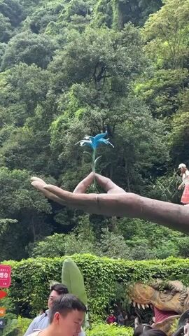 A video shows a person standing on a large sculpture of a hand. The hand is holding a blue flower. The person is wearing a white shirt, a red and white skirt, and a white hat. The person walks across the hand and then raises their arm. In the background, there are trees and a lush green hillside. In the foreground, there are people and a dinosaur sculpture. The video is set outdoors.