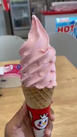 A person holds a pink soft serve ice cream cone in a waffle cone with a red wrapper featuring a snowman cartoon. The background shows a brightly lit store with food preparation and storage equipment. A sign reads "HOT DAY ICED TEA". The person is holding the cone, which is slowly rotating.
