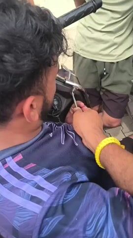 The video shows a close-up of a man's back and shoulder as he is bent over, appearing to work on the handlebars of a scooter. He is wearing a dark blue t-shirt with purple and white stripes. Another person, partially visible, is wearing a light green t-shirt and dark shorts, and is also looking at the scooter's handlebars. Two other individuals with curly dark hair are leaning in from the right side, also seemingly focused on the same area of the scooter. The man in the foreground has a yellow bracelet on his right wrist and a thin silver chain around his neck. The background is blurred but suggests an outdoor setting. The action appears to be an attempt to repair or adjust the scooter.