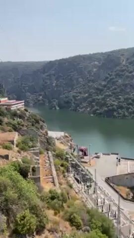 A slow, panning shot reveals a dam and reservoir in a mountainous region. The water is a pale green color. On the left is a red and white building on a series of terraces, along with electrical towers and equipment. The dam extends across the water, and a road runs on top of the dam. The sky is bright and clear. The mountains are covered in trees and other vegetation.