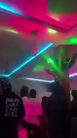 The video shows a group of people in a dimly lit, crowded room, possibly a party or club. The room is decorated with colorful lights, including pink, blue, and orange hues, and there are ceiling fans visible. Several individuals are dancing or moving to music. One person is wearing a black t-shirt with the words "Make That Risk" printed on the back. Another individual is wearing a white outfit. The atmosphere appears lively and social.