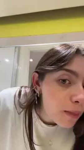 A young woman with fair skin and dark brown hair, wearing a white t-shirt and a necklace, is shown inserting an earring into her left ear. She faces a mirror with a yellow-green border. Her hair is parted and styled with loose waves. She is wearing several earrings in that ear already. The bathroom is visible in the reflection, with a frosted glass shower door visible. The woman's hands are visible, with a bracelet on her left wrist and pink polish on her nails. She is focused on the task of putting in the earring.