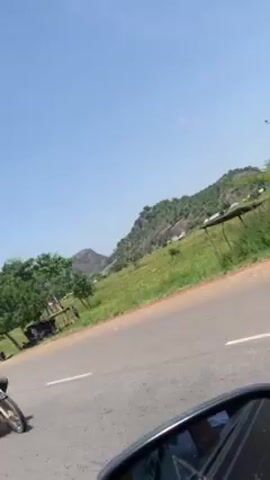 The video shows a roadside view from a moving vehicle, likely a motorcycle, as indicated by the side mirror visible in the bottom left corner of the frame. The camera is pointing towards a grassy field, dotted with trees and small structures resembling shelters or sheds. In the background, there are hills or small mountains covered with vegetation. The sky is clear and blue. The road is paved with a white line marking the edge. The focus is on the scenery passing by, and there is a strong wind noise audible in the recording.