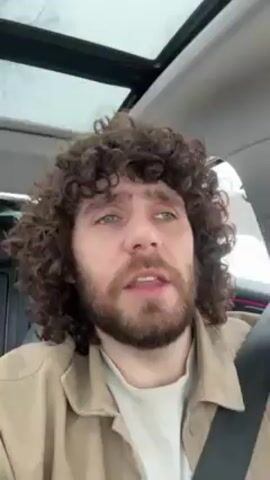The video shows a man with curly brown hair and a beard sitting in the driver's seat of a car. The car has a sunroof, which is visible in the background. The man is wearing a beige jacket over a white shirt. He is looking at the camera and speaking. The background outside the car appears to have snow or slush on the ground. The man is wearing a seatbelt. The car's interior appears to be dark in color, with some pinkish-red light reflecting on the steering wheel. There is music playing in the background.