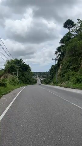The video displays a point-of-view shot of a paved road winding through a lush, tropical landscape. The road is an asphalt surface with white lane markings. On either side of the road, the terrain slopes upwards, covered in dense green vegetation and trees, including palm trees. Utility poles with wires run along the sides of the road.

Several vehicles are visible on the road. A white van with a black front is in the foreground, followed by a car and a motorcycle. Further down the road, more vehicles can be seen. The road appears to be leading towards a town or village nestled in the distance, with the sea visible on the horizon under a cloudy sky.

The weather appears to be overcast with large, dramatic clouds dominating the sky. The lighting suggests it is daytime, likely late morning or early afternoon. The presence of abundant greenery and palm trees indicates a warm, humid climate, consistent with a tropical location like Seuneubok, Indonesia. The overall mood conveyed is one of travel and exploration, with the open road leading to an unknown destination. No distinct emotions are explicitly portrayed, but the journey forward suggests a sense of purpose or adventure.

There is some visible text: a yellow license plate on the white van reads "DL 3363 FN". A green sign on a utility pole on the left side of the road is partially visible, but the text is unreadable.