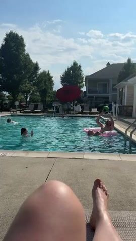 The video shows a poolside scene on a sunny day. In the foreground, a person's legs and feet are visible, suggesting they are lying down on a lounge chair. The middle ground features a swimming pool with several people. Two individuals are actively engaged in the pool: one person is in the water, reaching up with their arms, and another person is on a pink inflatable ring, also with their arms raised, appearing to be playing or interacting with someone off-camera. A green ball is visible in the water, indicating a game is in progress. In the background, there are trees, a building that appears to be an apartment complex or hotel, lounge chairs, and a red umbrella. The sky is blue with scattered clouds. The overall atmosphere is one of recreation and leisure.