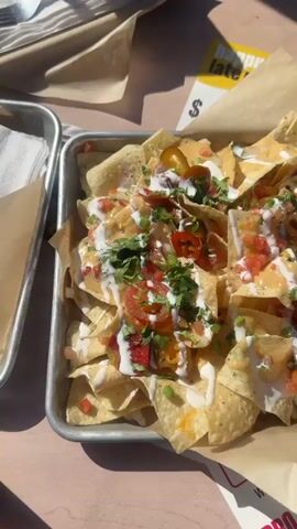 A high angle shot shows a tray of nachos on a wooden table, bathed in bright sunlight. The nachos, piled high on a metal tray lined with brown paper, consist of tortilla chips covered in cheese sauce, pico de gallo, jalapenos, a white creamy sauce, and chopped cilantro. A small black bowl of red salsa sits beside the nachos. A yellow card with the words "happy late hour" is visible in the background. Part of another tray with napkins is visible in the upper left corner. The table appears to be outdoors, with a metal railing casting a shadow on the right side of the shot.