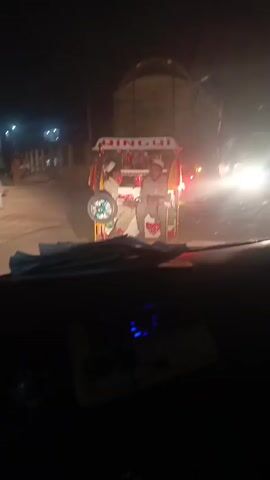 From the perspective of someone inside a car at night, the video shows a brightly lit auto rickshaw ahead with the name "QINGQI" displayed on top. The rickshaw has at least two passengers. There is a large truck parked nearby. The road appears dusty, and there are other people and vehicles in the background. The dashboard of the car is visible, with the speedometer indicating a low speed or standstill. The video appears to be taken in an area with minimal lighting.
