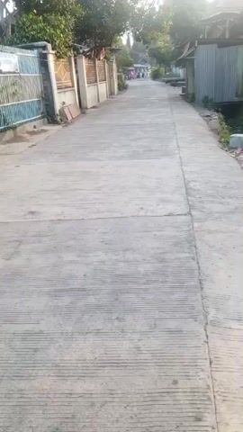 The video shows a concrete road in a rural or suburban area. There are houses and fences on both sides of the road. In the distance, a few people can be seen walking. The sun is shining, casting long shadows. The road appears to be in good condition, with some cracks and expansion joints visible. There is some vegetation growing along the sides of the road and in front of the houses.