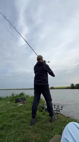 A man in a dark hoodie and black sweatpants is fishing near a lake. He is holding a fishing rod and appears to be casting. The lake is calm, with a few trees and a field in the background. The sky is cloudy. There are fishing rods set up on the bank.