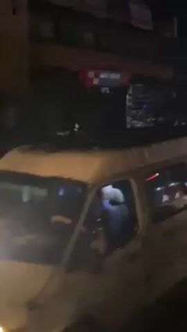 At night, a white van with a partially visible driver is parked near a building with an awning. People are walking on the street. A red car is parked beside the van, and people are interacting near the vehicles. A man is wearing a red shirt.