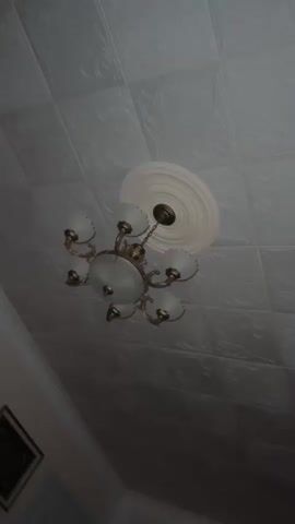 A video shows a chandelier hanging from a textured ceiling. The chandelier has multiple frosted glass shades and a brass-colored metal frame. The ceiling tiles have a raised floral pattern. A decorative ceiling medallion is visible around the base of the chandelier. The lighting in the room is dim, casting shadows on the ceiling. The camera angle is from below, looking up at the chandelier and ceiling. There is a hint of a framed picture on the wall in the lower left corner of the frame.