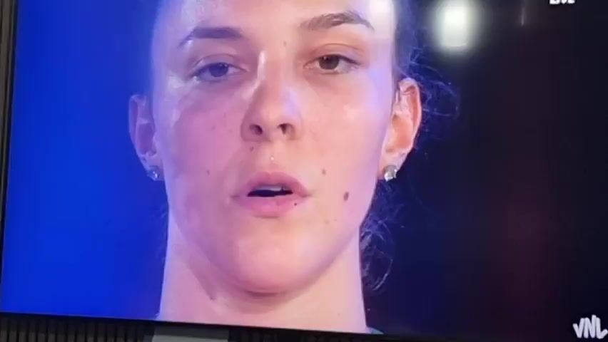 The video displays scenes from a women's volleyball event, likely taking place in an indoor arena in Xàtiva, Spain.

The initial shot is a close-up of a young woman's face. She has dark hair pulled back, light brown eyes, and a neutral, focused expression. Small stud earrings are visible, and she has a small mole below her lower lip and on her right cheek. The background is a blurred dark blue/purple. Visible text on the screen includes "VNL" in the bottom right corner and a partially visible "LIVE" in the top right.

The scene then widens to show a line of female athletes, presumably a volleyball team. They are standing side-by-side in an indoor sports arena. They wear matching red sleeveless jerseys with green trim around the V-neck and armholes. Various sponsor logos, such as "efbet" and "Lidl," are visible on their jerseys, along with player numbers like 20, 2, and 23. Their expressions are serious and attentive, with one player appearing to sing or having her mouth slightly open.

The setting is a well-lit indoor sports facility, with court markings and a net faintly visible in the background. A large screen or banner behind the athletes shows the partial word "GARIA," likely part of "BULGARIA." The activity appears to be a formal pre-game ceremony, possibly a national anthem presentation or team introduction, consistent with a professional sports match, specifically within the Volleyball Nations League (VNL) given the on-screen text.