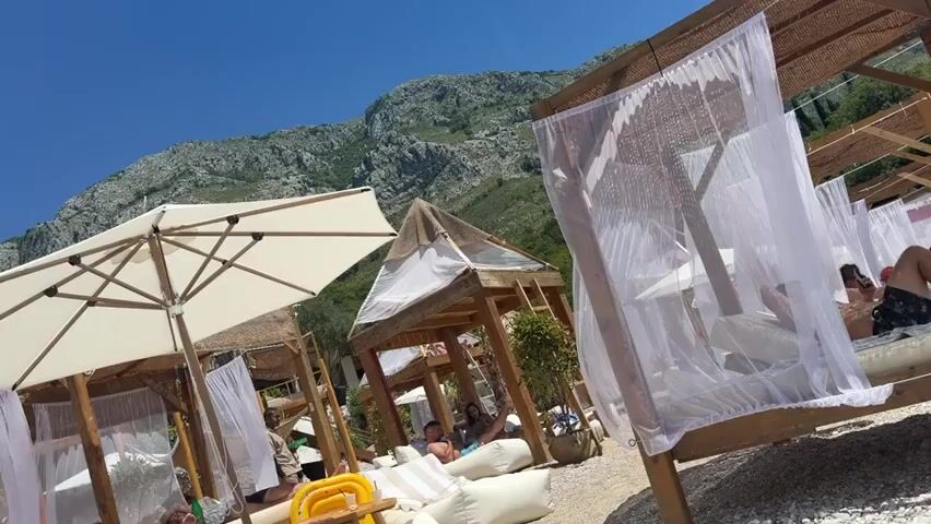 This is a horizontal, medium-shot video of a beach with several cabanas, umbrellas, and people enjoying a sunny day. The cabanas are made of wood and covered with white sheer fabric, creating private lounging areas. Inside some of the cabanas, people are seen relaxing on large cushions. There are also several large white umbrellas providing shade over the beach. In the background, there are mountains and the blue sky. The beach appears to be made of pebbles. The video is slightly tilted. There are at least 5 people visible in the video, either lounging in the cabanas or walking on the beach. The overall atmosphere appears to be relaxed and vacation-like.