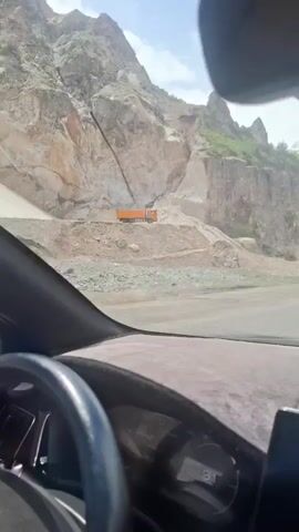 The video shows a view from inside a car driving through a mountain landscape. In the first part of the video, the car passes a quarry with a large orange truck parked in it. The view pans up to reveal snow-capped mountains in the distance, and a small creek running alongside the road. The scene is bright and sunny. No people are visible.