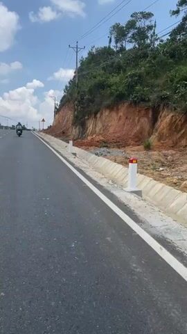 A motorcycle is driving on a road running alongside a steep slope with red-toned soil. The road is asphalt and features a white line marking the edge. A concrete drainage ditch separates the road from the base of the slope. There's a warning sign to the side of the road. The sky is partly cloudy, and overhead power lines stretch along the side of the road. Trees top the slope.