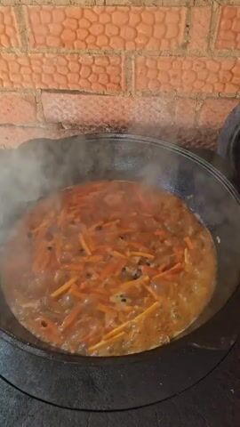 A large black cooking pot, possibly a kazan, is depicted from an overhead angle, resting on a black stovetop surface. Inside the pot, an orange-brown liquid containing numerous julienned carrots and darker fragments is vigorously boiling, releasing a substantial amount of white steam that rises from the surface. A reddish-brown brick wall with a textured pattern forms the background.