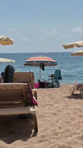 The video shows a sunny day at the beach with clear blue skies and calm ocean water. Several beach chairs, some with umbrellas, are set up on the sandy shore. In the foreground, a tan beach chair is empty. Further back, a colorful striped umbrella provides shade for a person lying down on a beach chair, along with some bags and a towel. Another beach chair is visible to the right, with a person lying on it. The water appears tranquil, with a few small waves. The overall atmosphere is relaxed and leisurely, typical of a beach vacation.