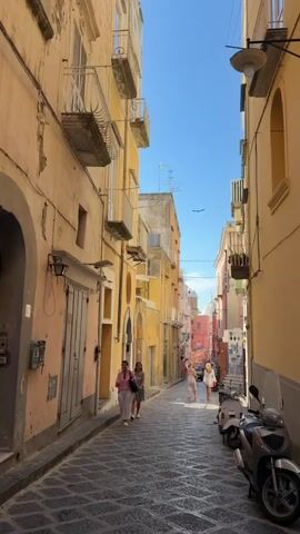 The video shows a narrow street in an old town with colorful buildings on both sides. The street is paved with dark cobblestones, and there are a few people walking down the street, including two women who stop to take pictures with their phones. Parked on the right side of the street is a silver scooter. The buildings are mostly pastel colors, with balconies and some decorative details. At the end of the street, a dome-shaped structure can be seen in the distance. The sky is clear and blue.