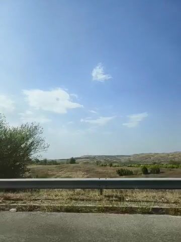 A video captures a view from a moving vehicle, likely a car. The foreground shows the edge of a paved road and a metal guardrail. Beyond the guardrail, a dry, grassy field stretches out, dotted with sparse trees and low hills in the distance. The sky is a bright blue with a few wispy white clouds. The sun appears to be high, indicating daytime. The overall impression is that of a scenic drive through a rural, possibly arid, landscape.