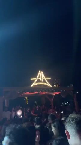 A lively crowd is gathered at an outdoor event at night. A neon sign with the letters "AA" in a stylized font is prominently displayed above a stage area. The stage is illuminated with a series of bright, circular lights and some vertical orange lights. There are people on the stage, some appearing to be performers or DJs, interacting with the crowd. The atmosphere is energetic, with smoke or fog machines creating a hazy effect, catching the stage lights. The sky above is dark, with some faint lights visible in the distance, possibly from other structures or a city skyline. The overall impression is of a nighttime concert or party scene.