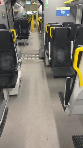 The video shows the interior of a train car. The seats are black with a white polka-dot pattern and yellow accents on the headrests. The floor is gray and there are metal treads between seating areas. A display shows the name of a stop, "Riga," and an advertisement for public transportation. A woman can be seen in the reflection of a train window.