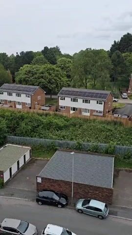 The video features an overhead view of a residential area, panning smoothly from left to right. In the foreground, a street is lined with several small brick buildings, possibly garages, with grey roofs. Multiple cars are parked along the street, including black, light blue, and white vehicles. A street light pole stands near one of the buildings. Beyond the street and buildings, a dense area of overgrown green bushes and vegetation is visible, bordered by a metal fence. In the midground and background, several two-story houses with white exteriors and dark roofs are visible, many equipped with solar panels. Green trees and dense foliage form a backdrop behind the houses, extending towards the horizon under an overcast sky. No people are visible.