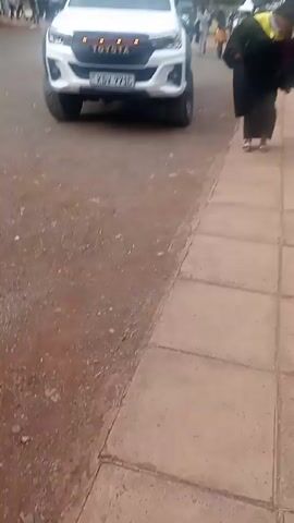 The video shows a person in a graduation gown walking on a paved path. A white Toyota truck is parked nearby. The camera follows the person as they walk toward a gathering of people in a park-like setting. Several other people are walking along the path, some also wearing graduation gowns. Green bushes line the path.