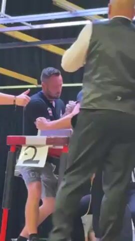 Here is a description of the video:

The video shows two men engaged in an arm-wrestling competition on a red and white table, marked with the number "2". They are both gripping hands tightly, facing each other, with a referee in a striped shirt standing on the side. Other men are observing the match. The man in a black shirt and grey shorts is on the left side of the table, and the man in a black shirt and black pants with a cap is on the right side. A man in a vest and pants is standing in the background watching the match. There is a sign and a grid of lights above the table.