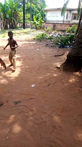The video captures an outdoor scene in Akwa Etiti, Nigeria, under bright daylight. The ground is covered in reddish-brown sand.

At the beginning (0:00-0:02), two young men are visible. One, shirtless and wearing light-colored trousers, stands facing the camera. The other, shirtless and clad in red shorts, stands with his back partially to the camera. The background features dense tropical vegetation, including palm trees and banana plants, and a light-colored building. Some scattered debris and trash are visible on the ground and amidst the foliage.

From 0:03, the focus shifts to a small, shirtless boy wearing light-colored pants, which have the word "GOOD" printed on them. He is being held and lowered towards the ground by an unseen individual whose hands are on his back. The feet of another person, wearing blue pants and sandals (one black-and-white striped, one plain black), are seen next to the boy. The boy appears to be struggling or resisting as he is lowered, arching his back. A brown husk (possibly a coconut) and a black case are also visible on the ground nearby. The scene suggests a playful or perhaps mildly coercive interaction involving the children.